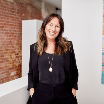 Professional indoor portrait of a smiling woman with long brown hair, wearing a black outfit and pendant necklace, standing against a brick and white wall background.